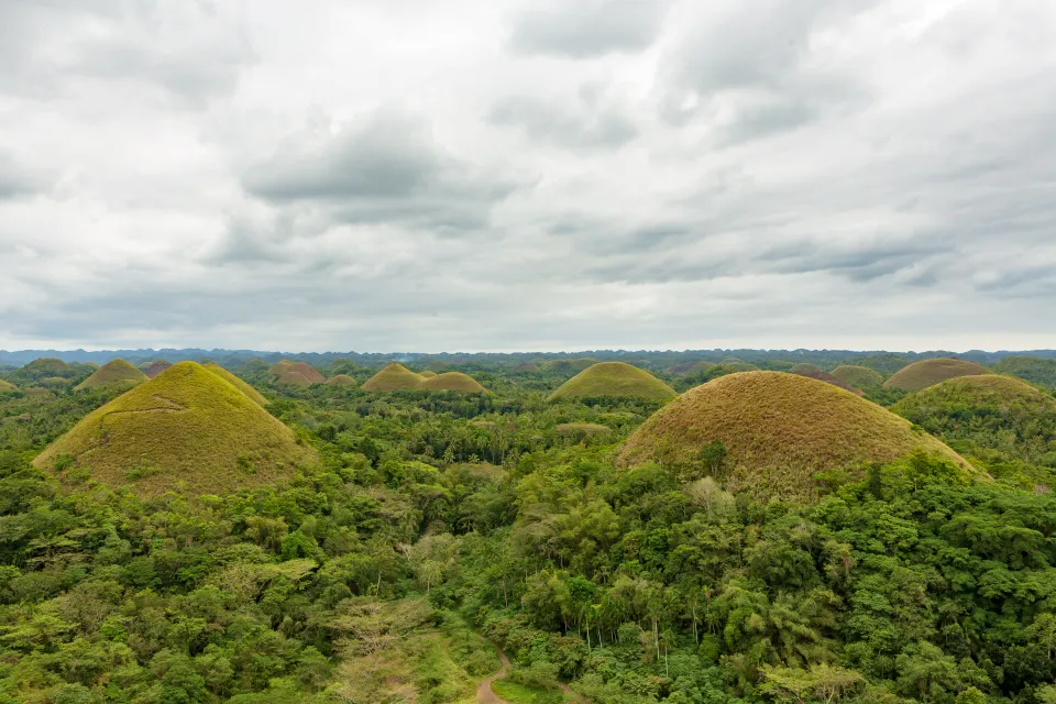 Pohled na Čokoládové kopce (Chocolate Hills) na ostrově Bohol, Filipíny. Ikonická krajina s desítkami symetrických, travnatých kopců obklopených bujnou tropickou vegetací pod zataženou oblohou.