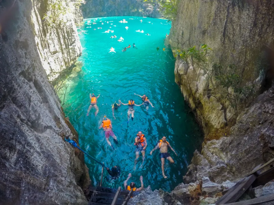 Turisté plavou v tyrkysové vodě Twin Lagoon poblíž ostrova Coron na Filipínách. Strmé vápencové útesy obklopují úzký vstup do laguny, zatímco sluneční světlo proniká vodní hladinou a vytváří oslnivé odrazy. Někteří návštěvníci nosí oranžové záchranné vesty, zatímco jiní volně plavou v klidné, průzračné vodě. Dřevěné schody a lano připevněné ke skalní stěně slouží jako vstupní bod do této malebné přírodní atrakce.