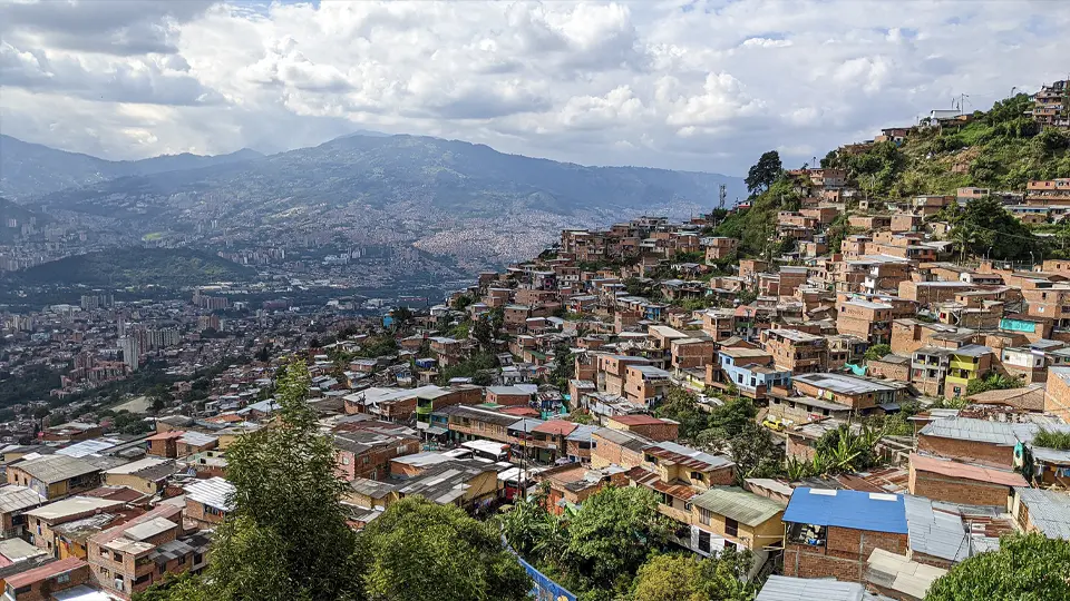 Panoramatický pohled na město Medellín v Kolumbii, pořízený ze svahu v jedné z městských chudinských čtvrtí (slumů); v popředí jsou hustě natěsněné cihlové domky s plechovými střechami rozeseté po zeleném kopci, v pozadí se rozkládá moderní městská zástavba v údolí obklopeném horami, pod částečně zataženou oblohou.