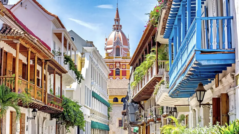 Barevná ulice v historickém centru Cartageny v Kolumbii, lemovaná koloniálními budovami s dřevěnými balkony a květinami, s dominantní věží katedrály svatého Petra Klavera v pozadí pod jasnou modrou oblohou.