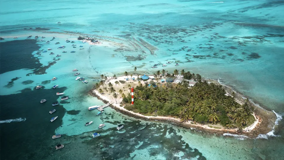 Letecký pohled na malý tropický ostrov Rocky Cay u pobřeží San Andrés v Kolumbii, obklopený tyrkysově modrou vodou a korálovými útesy, s bílou písečnou pláží, palmami, červenobílým majákem a množstvím lodí kotvících v okolí.