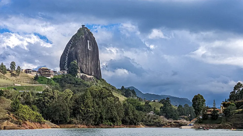 Pohled na monumentální žulový monolit El Peñol v Guatapé v Kolumbii, tyčící se nad zelenou krajinou s okolními budovami a lesy, pod zataženou oblohou, s vodní hladinou přehrady v popředí.