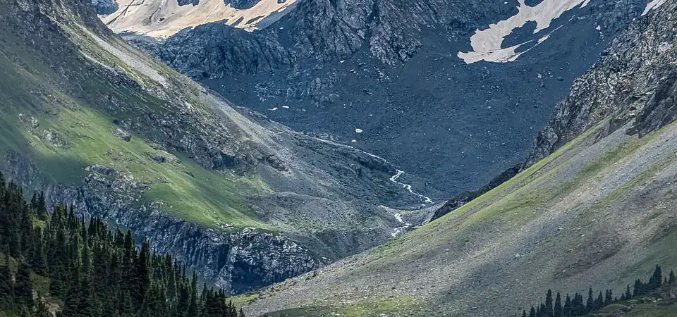 Horská krajina s impozantním zasněženým vrcholem v pozadí, obklopená strmými skalnatými stěnami a zelenými svahy. V popředí protéká křišťálově čistá horská řeka, obklopená trávnatými loukami a rozházenými kameny. Na svazích rostou husté jehličnaté lesy, které dodávají krajině dramatický kontrast. Částečně zatažená obloha vytváří dynamickou atmosféru se hrou světla a stínu.