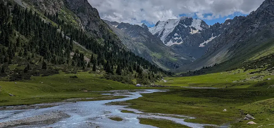 Malebné horské údolí s křišťálově čistou řekou, která se vine přes zelené louky. Řeka má několik menších ramínek a plytčin, přičemž voda odráží světlo z oblohy. Na svazích údolí jsou husté jehličnaté lesy a strmé skalnaté hory. V pozadí se tyčí zasněžené vrcholy. Obloha je poseta bílými mraky s jasně modrými úseky, což vytváří klidnou a přírodní atmosféru.