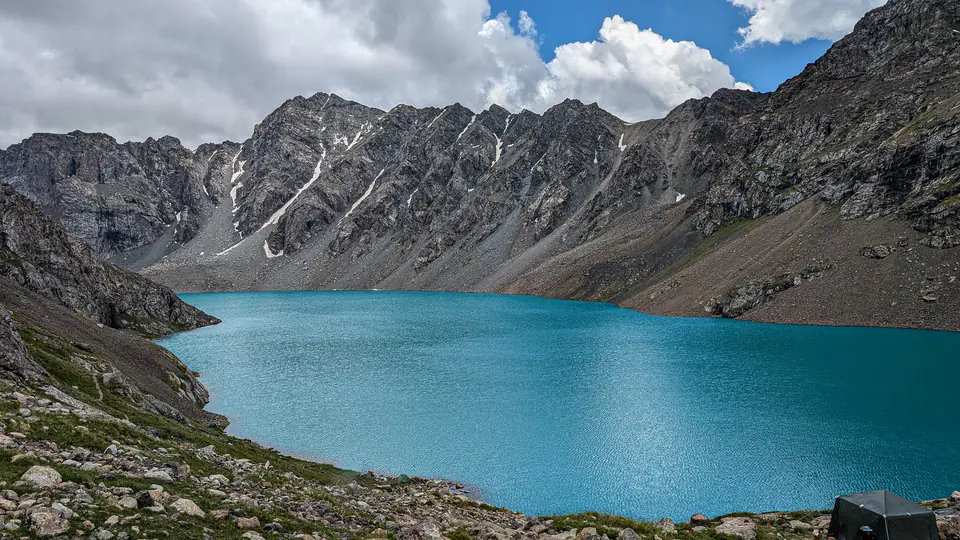 Tyrkysové jezero Ala-Kul ležící v dramatickém horském údolí Kyrgyzstánu, obklopené skalnatými štíty se stopami sněhu. Husté mraky se vznášejí nad horami, odrážející se na klidné vodní hladině. Tato scenérie je typická pro odlehlé alpské oblasti Střední Asie.