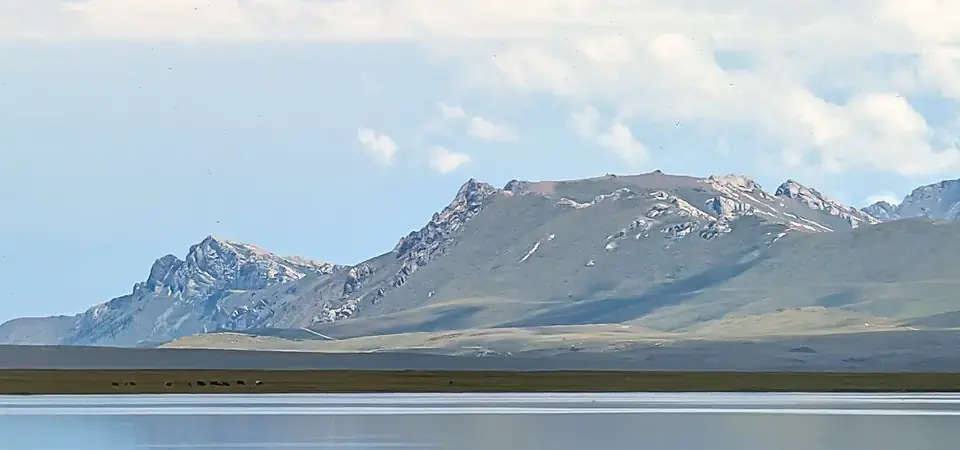 Osamělý šedý kůň pasoucí se na břehu jezera Son-Kul v Kyrgyzstánu. Klidná hladina odráží oblohu, zatímco v dálce se tyčí majestátní hory. Atmosféra je poklidná a typická pro pastýřskou krajinu Kyrgyzstánu.