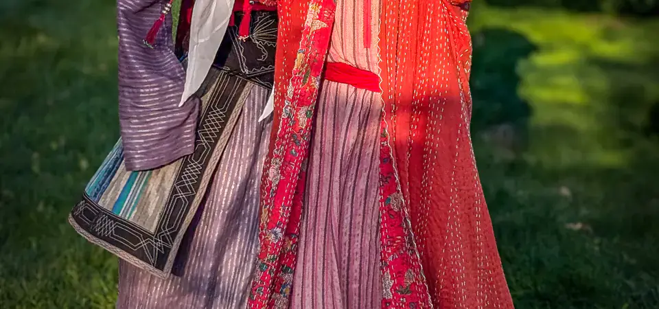 Dvě mladé dívky v bohatě zdobených kyrgyzských krojích s výraznými pokrývkami hlavy pózují v parku. Jedna z nich se usmívá a zvedá ruku před sluncem. Jejich oděvy jsou detailně vyšívané a reprezentují kulturní dědictví Kyrgyzstánu.